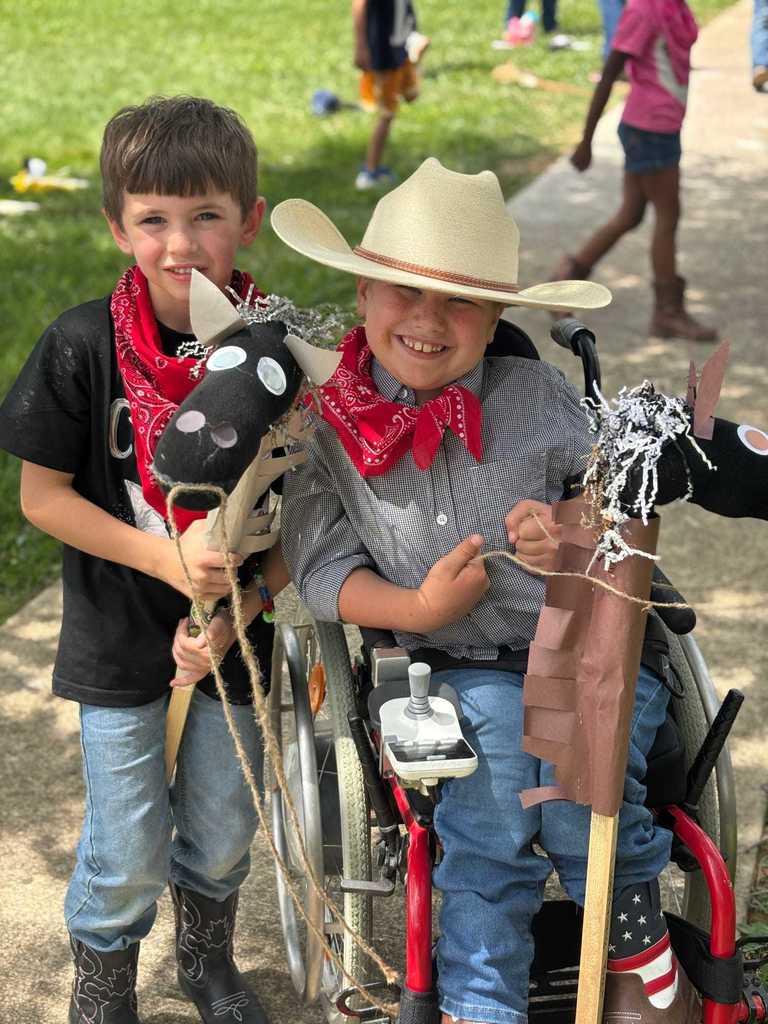 Two children dressed in cowboy-themed outfits, including red bandanas and cowboy hats, hold stick horses. One child is sitting in a wheelchair. In the background, other children are playing on a grassy area.