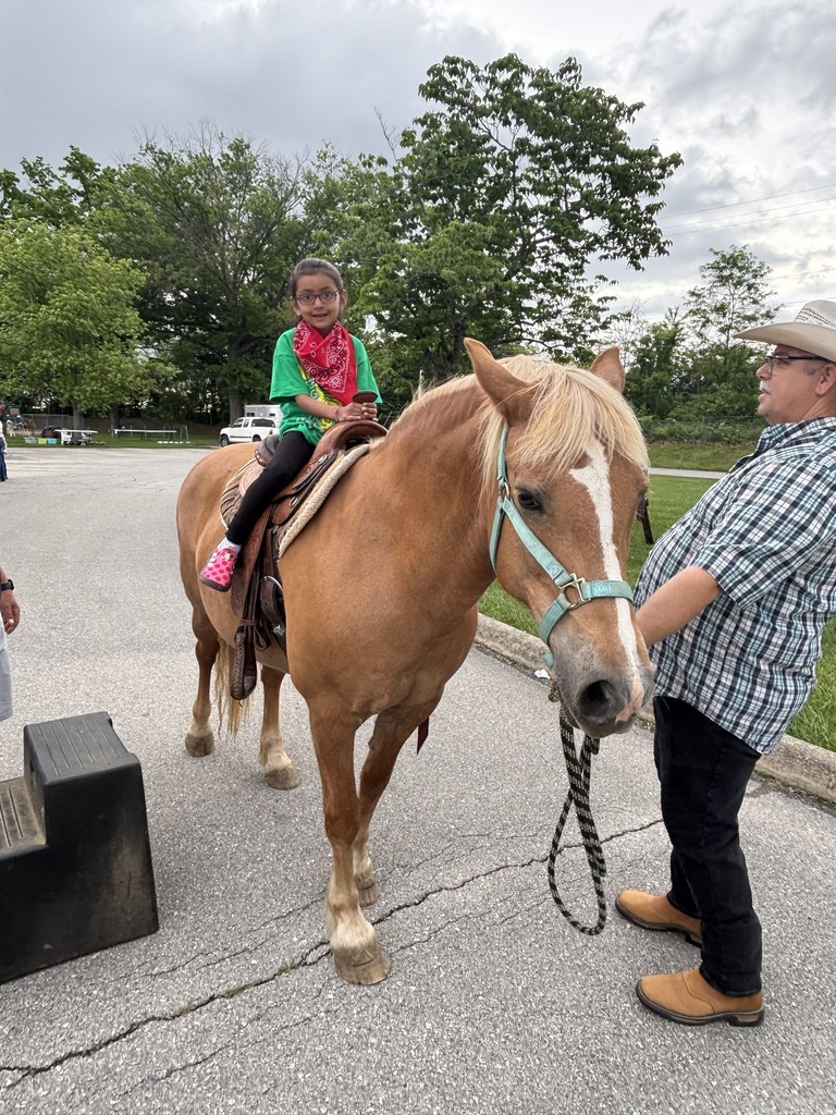 A child wearing a green shirt, red bandana, and black pants sits on a brown horse with a light mane. The horse is saddled and bridled, and an adult in a plaid shirt and jeans holds the reins. The scene is outdoors on a paved area with trees and grass in the background, and a black mounting block is positioned near the horse.