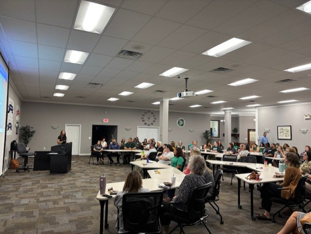 A modern, well-lit room with numerous people seated at tables, all facing a speaker standing near a podium at the front. A ceiling-mounted projector is visible, and the audience appears to be attentively engaged in the presentation.