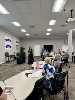 A group of people seated at tables in a conference room, facing a speaker at the front. The room is lit with fluorescent lights, and banners are visible on the walls.
