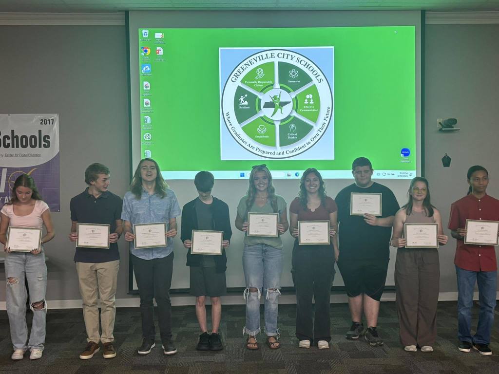 A group of nine individuals stand in a line holding certificates in front of a screen displaying the Greeneville City Schools logo and the motto "Motivating and Preparing Students for Lifelong Success." 
