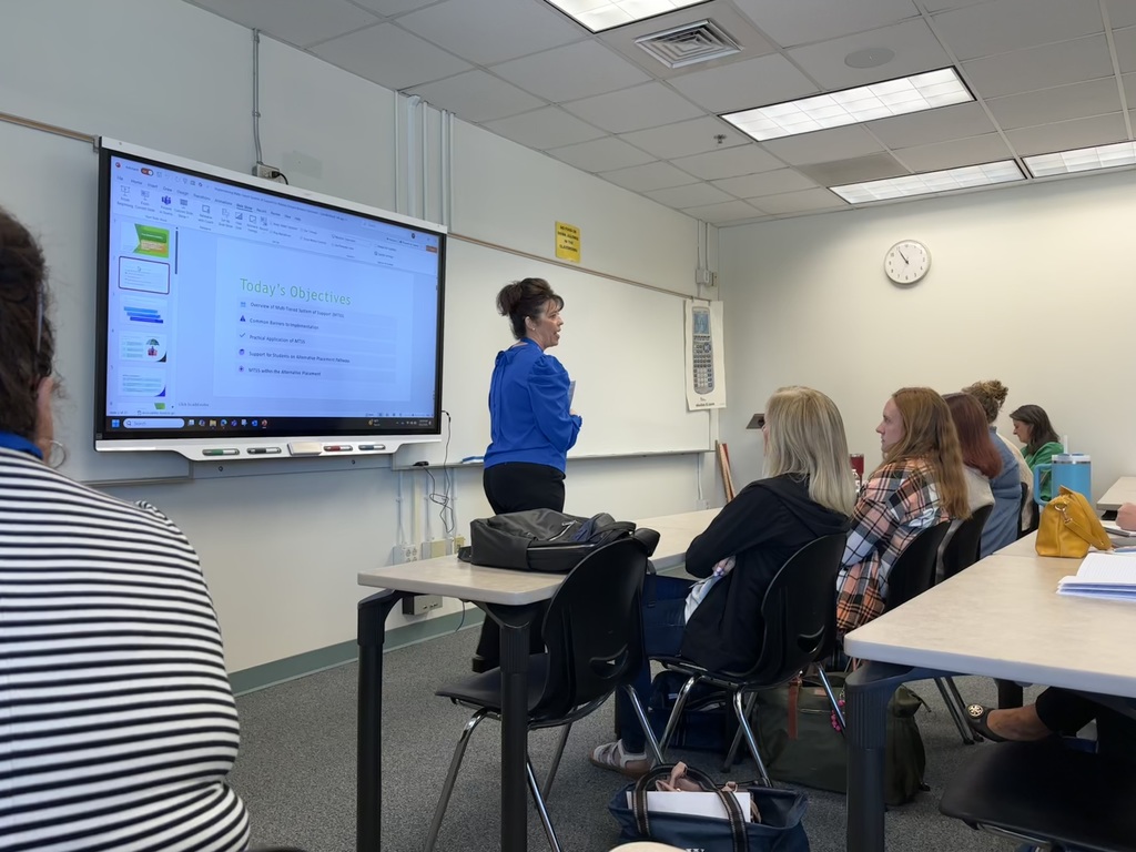 Classroom setting with several people seated at tables, facing a large screen displaying a presentation slide titled 'Today's Objectives.' A person stands near the screen. A wall clock shows the time as approximately 10:10