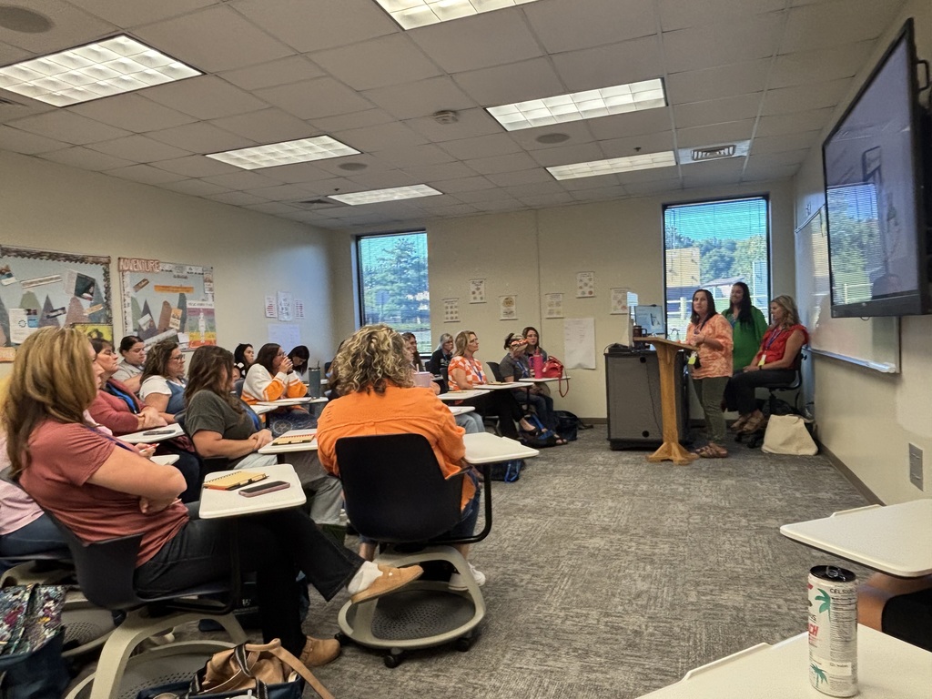 Classroom setting with several people seated at desks, watching a presentation by three individuals standing at the front. The room is lit with fluorescent lights and has posters on the walls.