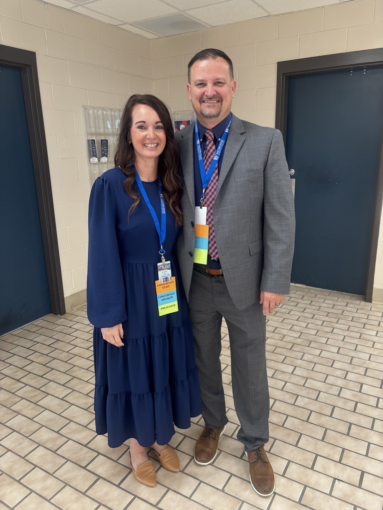 Two individuals standing side by side in a hallway with tiled flooring and blue doors. The person on the left wears a long blue dress and brown shoes, while the person on the right wears a grey suit with a tie and brown shoes. Both have lanyards with ID badges.