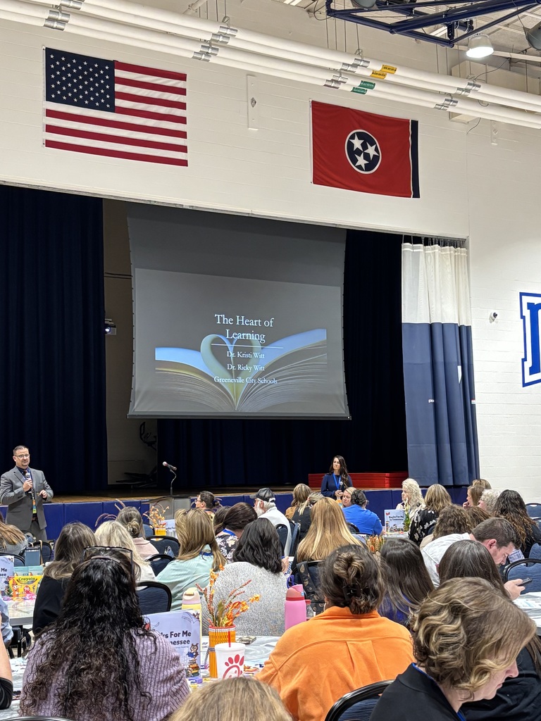 Large group of people seated in an auditorium or gymnasium, facing a stage with a speaker at a podium. A projection screen displays the text 'The Heart of Learning Dr. Kristi Witt Dr. Ricky Witt Greeneville City Schools' with an open book graphic and partially unreadable additional text.