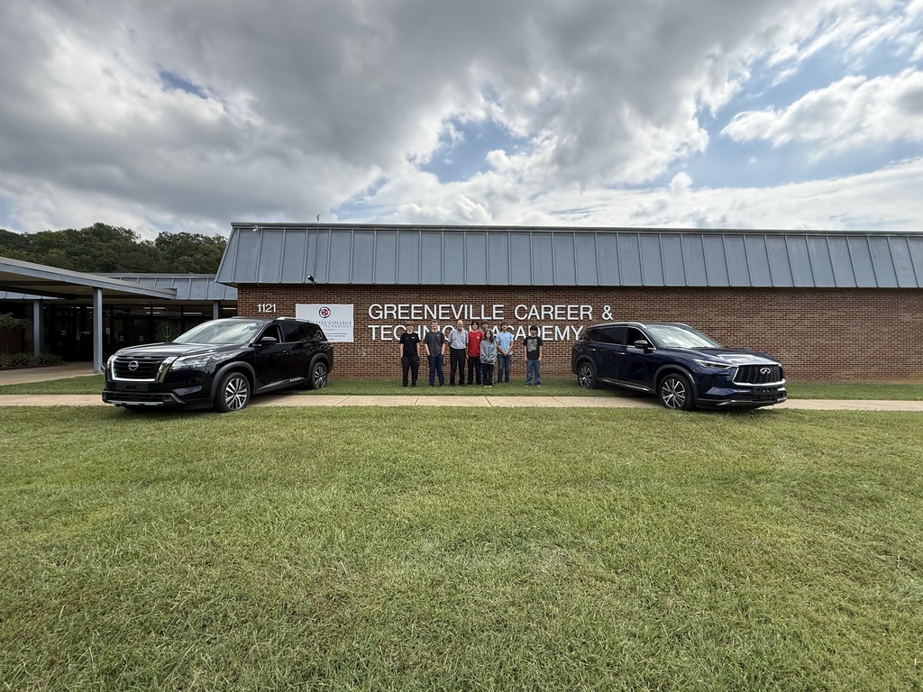 Group of people posing in front of the Greeneville Career & Technology Academy building, which has a brick facade and metal roof. Two black cars are parked on either side of the group. The sky is partly cloudy, and green grass is visible in the foreground