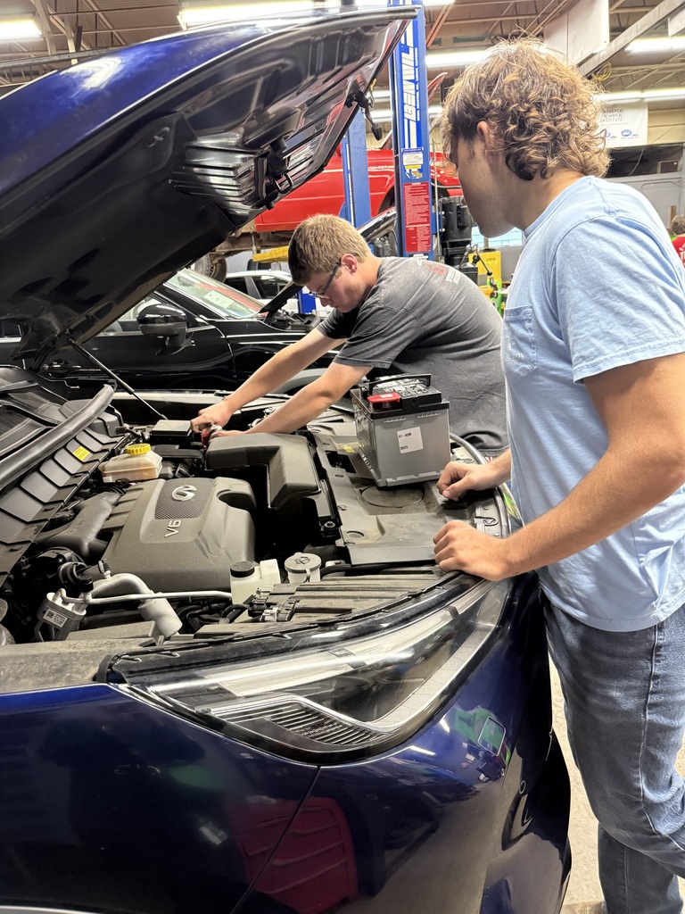 Two teenagers working on a car engine in an automotive repair shop. One holds a car battery while the other uses tools on the engine, which has a visible 'V6' marking. The open hood reveals the engine compartment, with automotive equipment and another vehicle in the background.