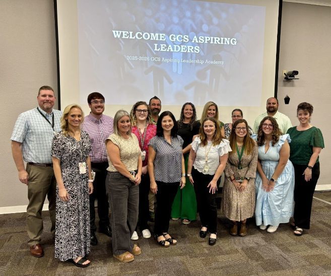 **Alt Text:** A group of educators standing together in a conference room in front of a large projection screen. The screen displays the text: “WELCOME GCS ASPIRING LEADERS – 2025-2026 GCS Aspiring Leadership Academy.” The individuals are arranged in two rows, with some standing and others slightly forward. The setting includes a neutral-colored wall, carpeted floor, and a mounted camera on the right side of the screen. The image represents a formal welcome event for participants in the leadership program.