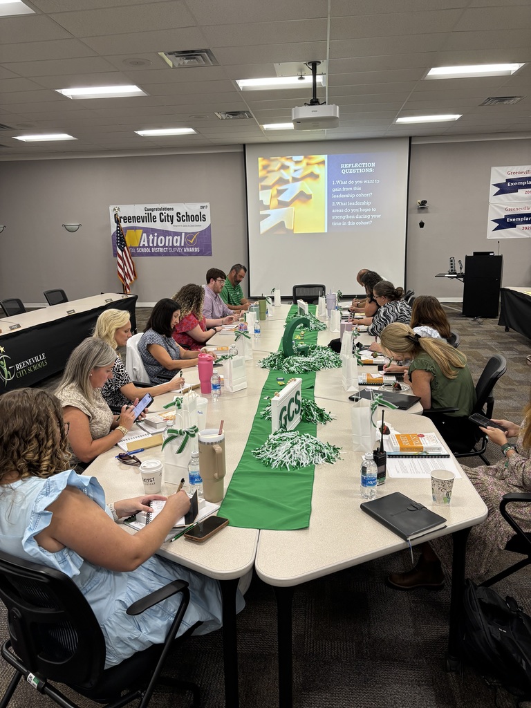 A group of educators seated around a large rectangular conference table in a meeting room. The table is decorated with a green table runner, pom-poms, and a centerpiece featuring the letters “GCS.” Participants have notebooks, papers, drinks, and electronic devices in front of them, actively engaged in writing or reading. In the background, a projector screen displays a slide titled “Expectations,” and banners on the walls read “Greeneville City Schools” and other motivational messages. The setting conveys a professional development session focused on leadership growth.