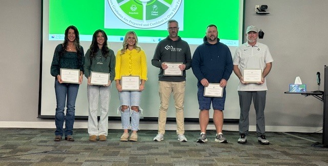 A group of six individuals stands in a line, each holding a certificate during an award or recognition ceremony. They are positioned inside a room featuring a large green screen or display behind them, which partially shows a circular logo or emblem. The individuals are dressed casually, suggesting a celebratory atmosphere.