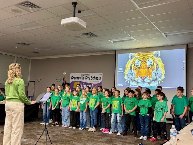 In a conference room setting, a group of children stands facing an adult woman who is conducting them. The children are wearing green T-shirts with text and images on the front. Behind them is a large projection screen displaying an image of a tiger's face, characterized by bright yellow and orange fur and piercing eyes. A banner reading "Congratulations Greenville City Schools " is also visible. The room features a ceiling with recessed lighting and has a water bottle on the table to the right.