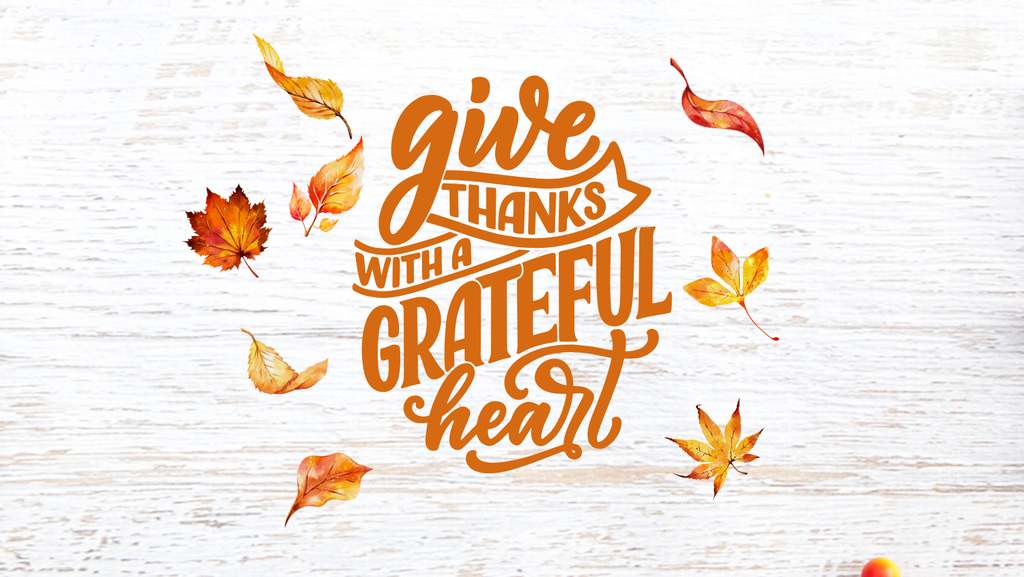White wooden background with scattered autumn leaves in shades of orange, yellow, and red. Centered in bold orange script text reads: ‘Give thanks with a grateful heart.’