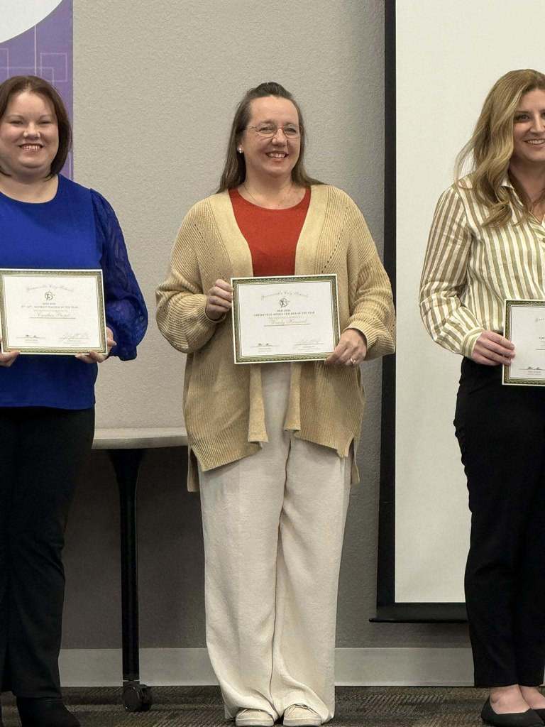 Three adults stand side by side indoors, each holding a framed certificate at chest height. The person on the left wears a bright blue top and dark pants. The person in the center wears a tan cardigan over a red top with light-colored pants. The person on the right wears a striped, long‑sleeve shirt and dark pants. A neutral-colored wall and a projection screen are visible in the background, suggesting a formal presentation or recognition event.