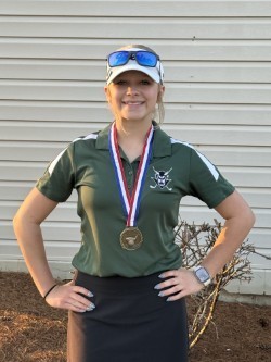 Person standing outdoors with hands on hips, wearing a green polo shirt, a baseball cap with sunglasses, and a medal on a red, white, and blue ribbon, in front of a light-colored building.