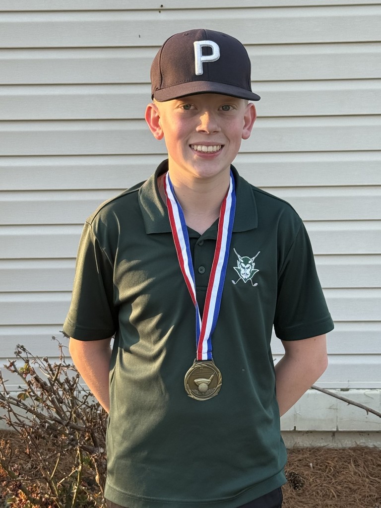 Person wearing a green polo shirt and a baseball cap with a “P” on the front, standing outdoors in front of a light-colored building, with a medal on a red, white, and blue ribbon around the neck.