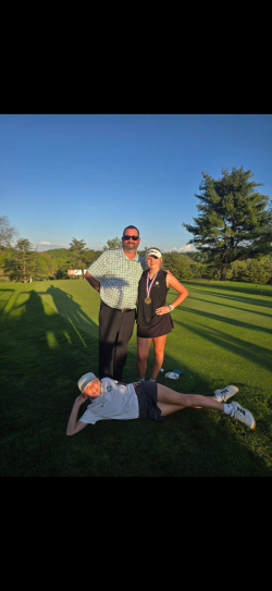 Three people pose on a golf course during golden‑hour lighting. Two individuals stand upright in the background near a putting green, while a third person lies playfully on the grass in the foreground, propped on one elbow. The standing individuals wear golf attire and medals around their necks. Trees and distant hills are visible under a clear sky.