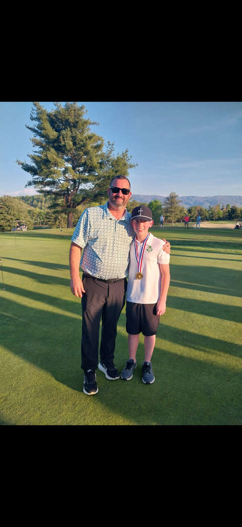 Two people stand side by side on a golf course, both wearing golf attire. One person wears a medal around their neck. The photo is taken on a well‑maintained green with trees and mountains visible in the background. 