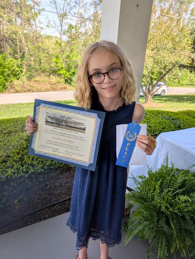  a student standing outdoors, holding a framed certificate in one hand and a blue first‑place ribbon in the other, surrounded by greenery and a porch setting.