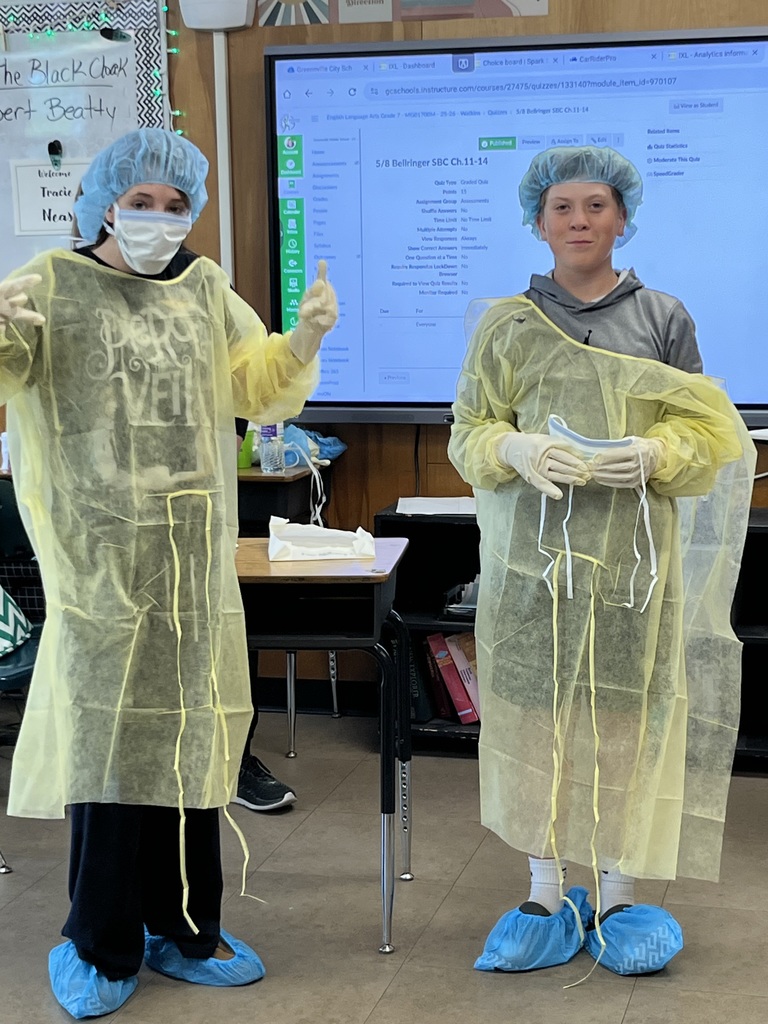  Two individuals stand side by side in a classroom wearing similar protective medical-style gowns, gloves, and hair coverings, demonstrating or posing with the equipment.