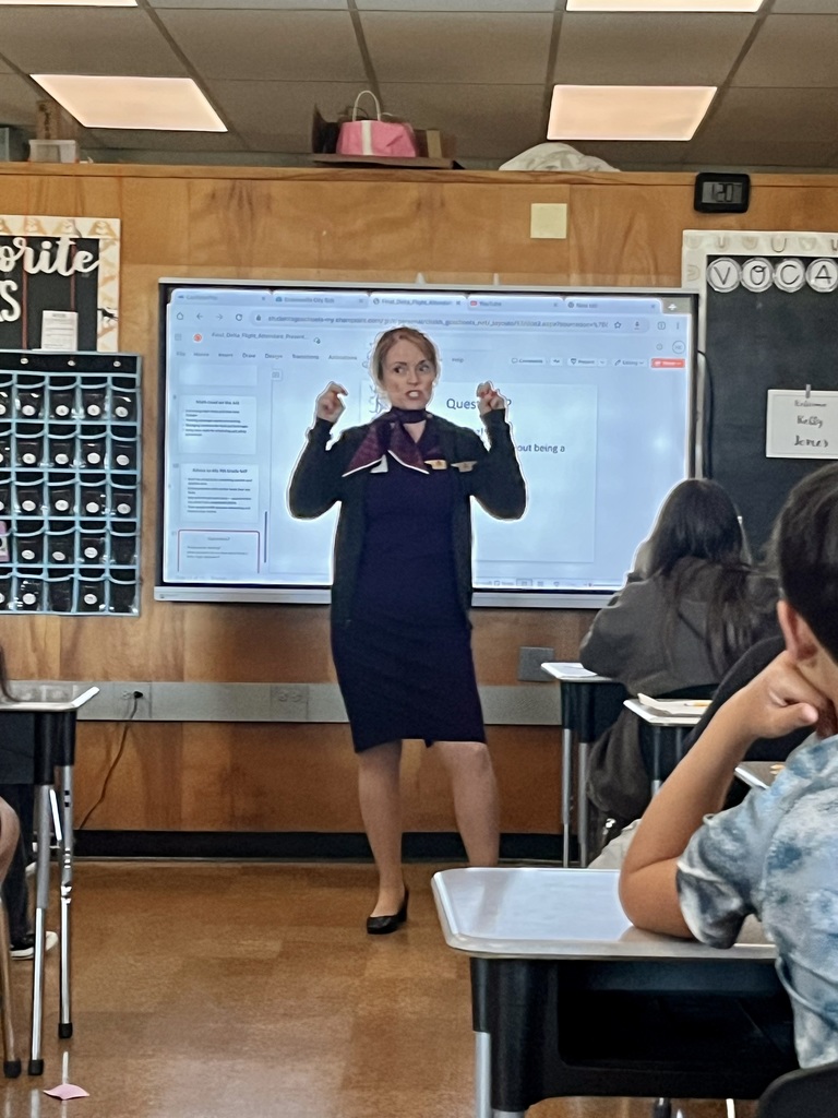A classroom with a digital display screen. An adult stands at the front demonstrating or speaking, while students sit at desks facing forward.
