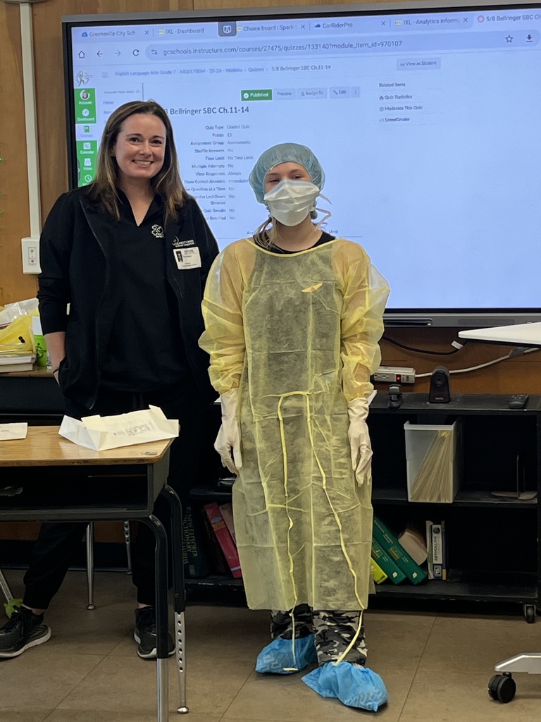 Two people stand in front of a classroom display screen. One person is wearing protective medical-style gear including a disposable gown, face mask, hair covering, and gloves. A desk with papers and supplies sits in the foreground.