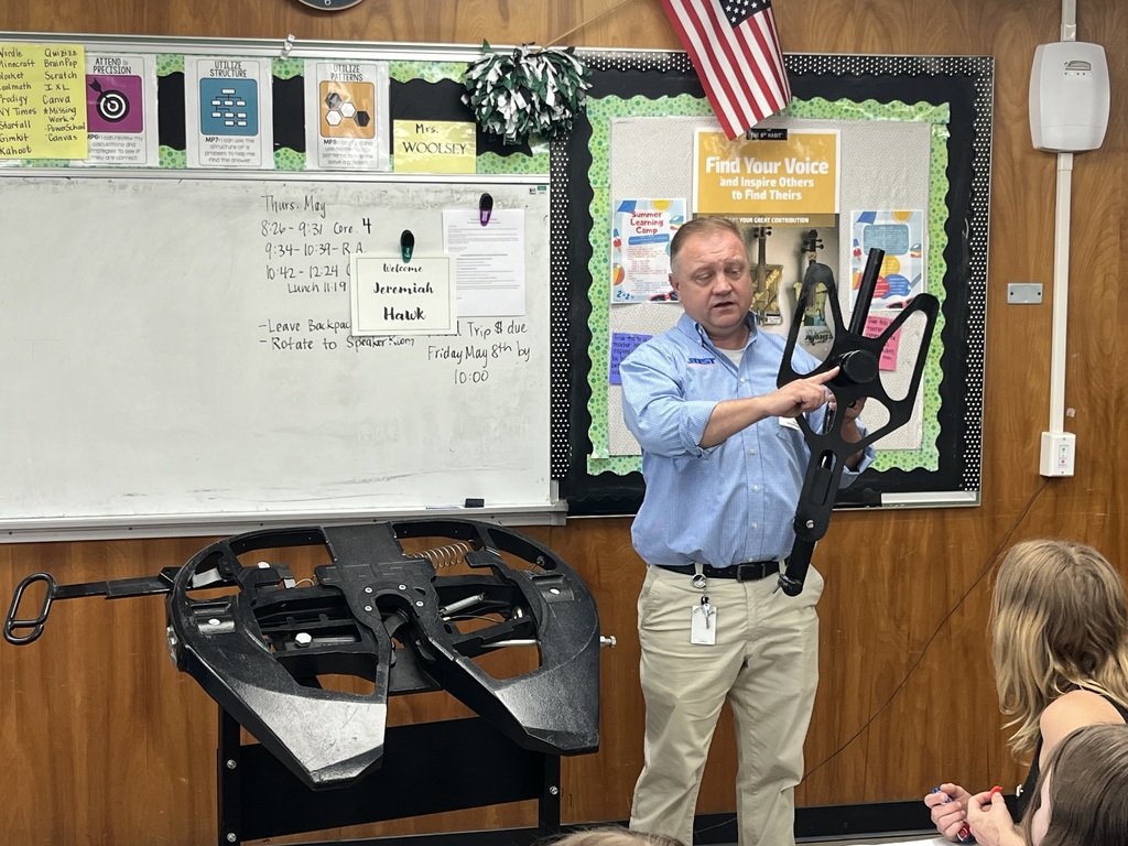  In another classroom setting with a whiteboard and posters, an adult holds up a large mechanical or drone-like component while explaining its features.