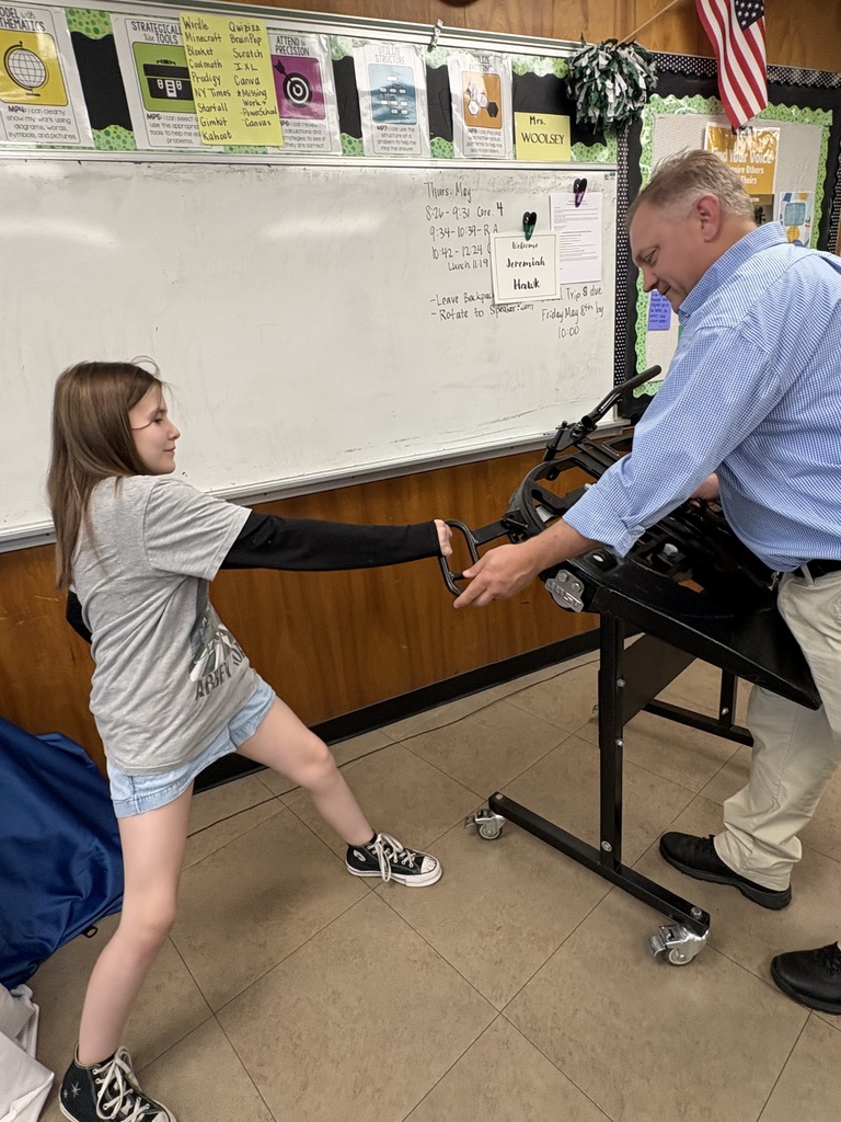 A classroom with a whiteboard in the background. An adult is demonstrating a piece of equipment to a student standing nearby, holding part of the device while the student extends an arm forward.