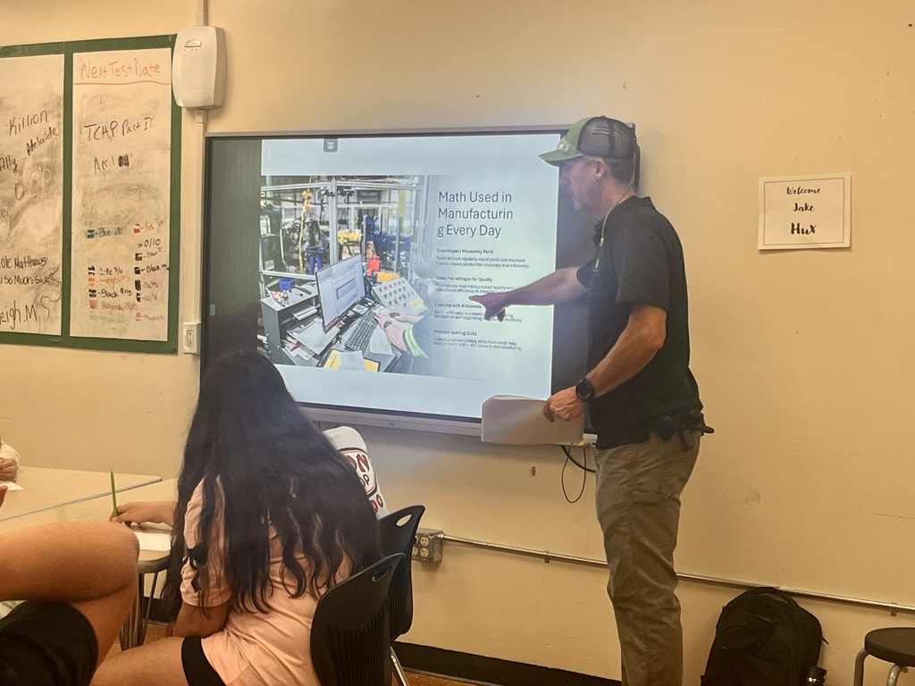  classroom with a digital display screen. An adult stands at the front demonstrating or speaking, while students sit at desks facing forward.