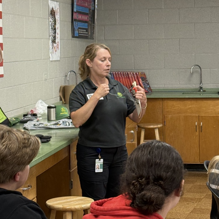 a woman in a gray t-shirt stands in front of a class of middle school students  