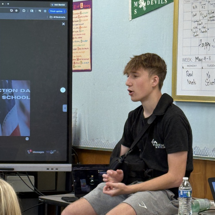 a man with an arm brace sits on a table while explaining his career to students.  on a table 