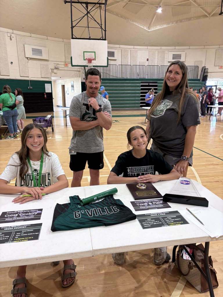 two adults stand smiling behind two children seated at a table with cross country and track & field papers on it.