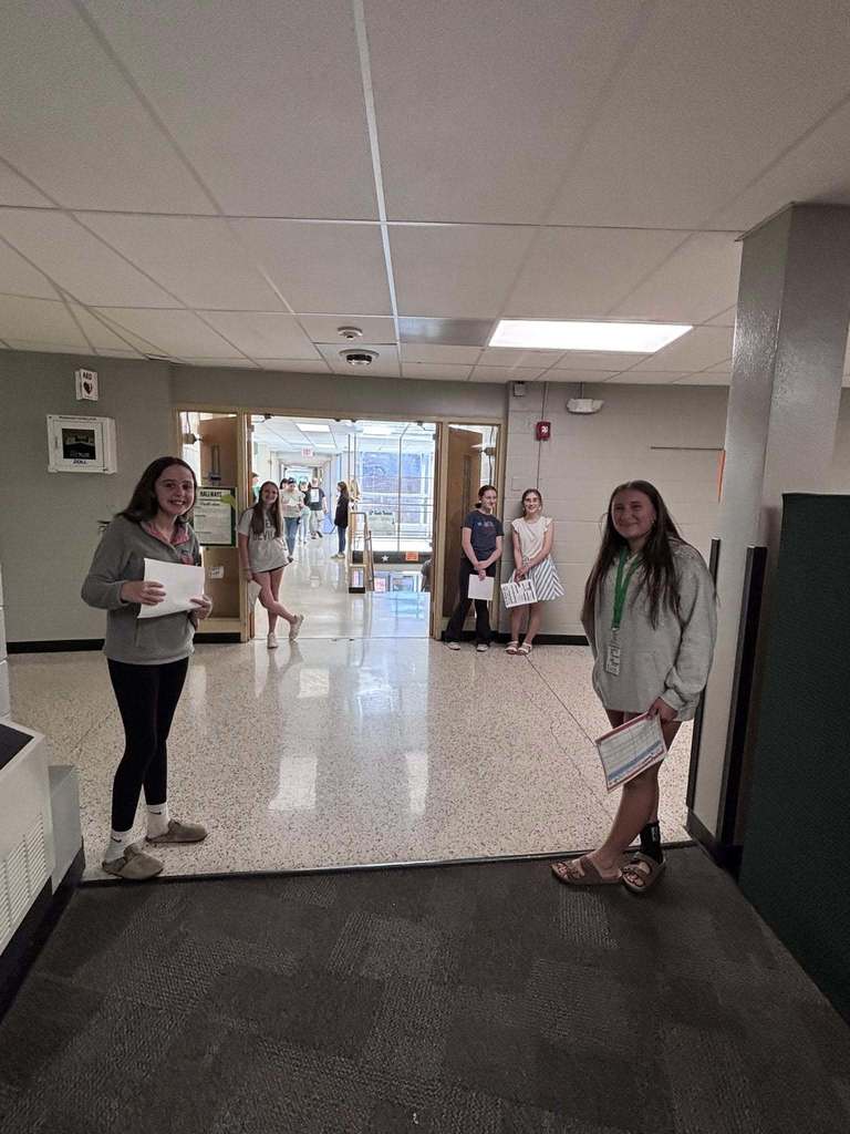 A school hallway with a tiled floor and drop ceiling is shown from a doorway. Several students are standing and facing the camera, each holding papers. One student stands in the foreground on the right near a wall and doorway, while another stands to the left holding a sheet of paper. In the middle background, more students are gathered near an open doorway that leads to a brighter corridor with lockers and natural light. The walls are painted neutral colors, and visible features include a ceiling light panel, a security camera, a fire alarm, and an AED cabinet mounted on the wall.