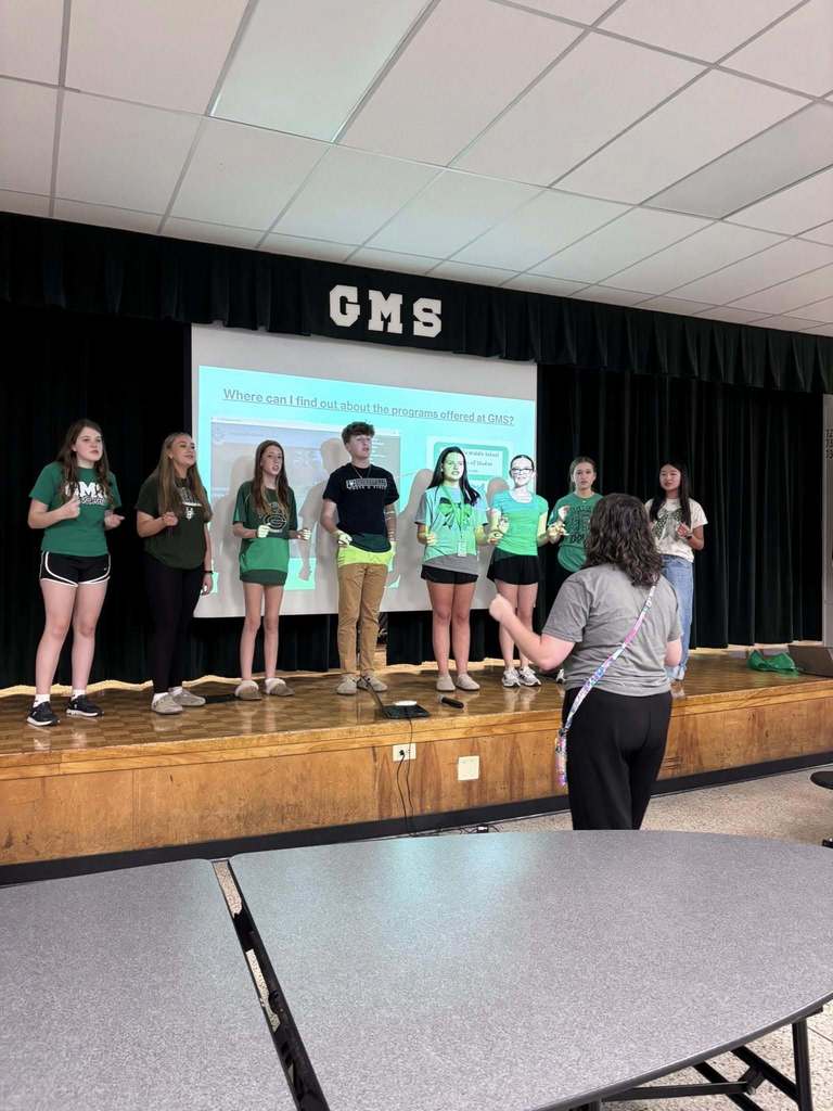 8 chorus students stand on a stage while a teacher instructs them
