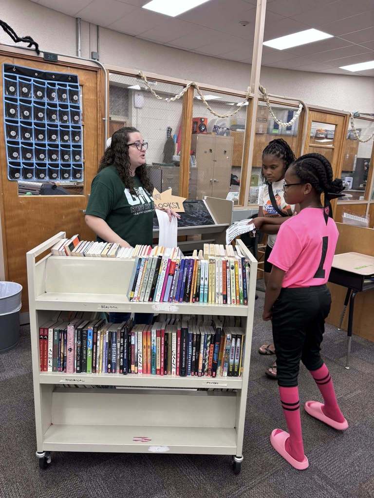 two students stand in a library looking at books while a teacher speaks to them.