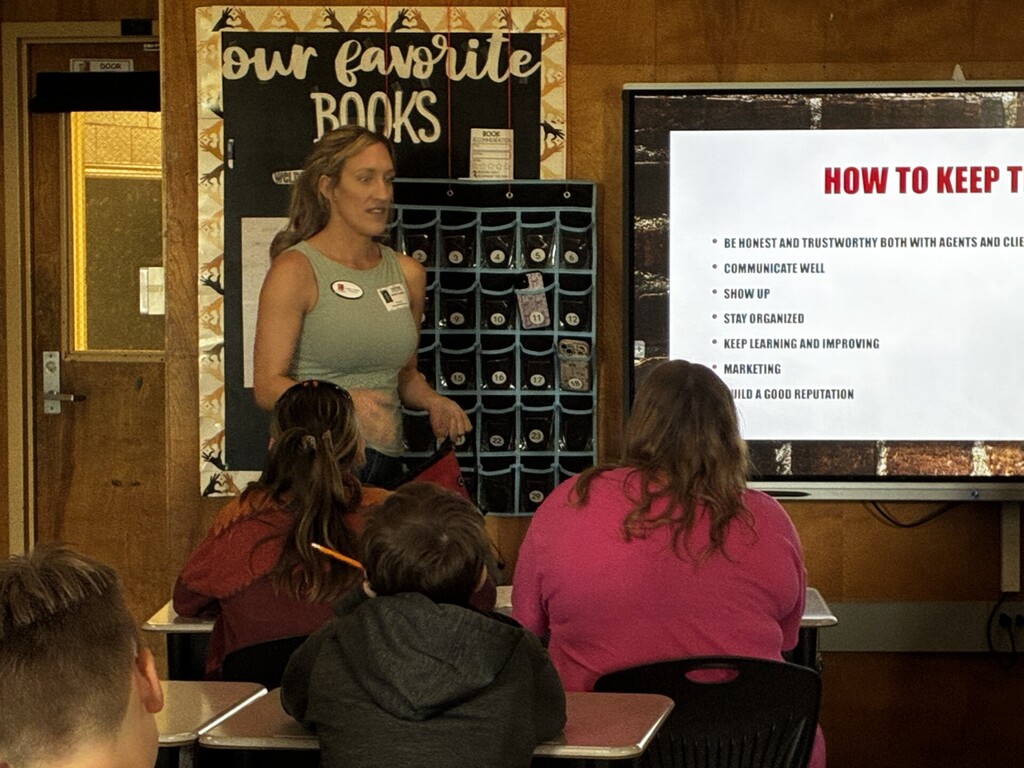A classroom presentation is displayed on a large screen with the title “How to Keep the Job?” and a list of workplace skills, including communication and organization. A presenter stands beside the screen while students sit at desks facing forward.