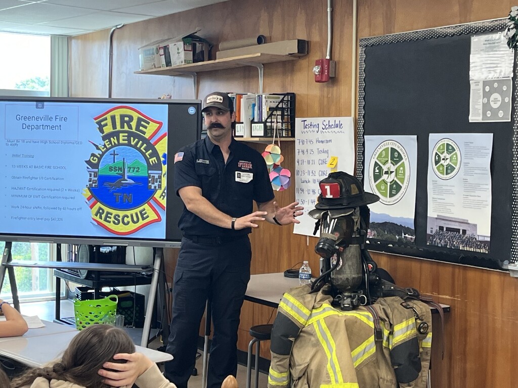 A fire department presenter stands at the front of a classroom speaking to students, with a display screen showing “Greeneville Fire Department” and a fire rescue logo. A full set of firefighter gear, including a helmet and jacket, is set up on a table beside the presenter.
