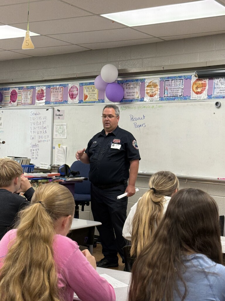 A community presenter in a fire department uniform speaks to a classroom of seated middle school students during Career Day. A whiteboard with a daily schedule and classroom materials is visible behind the speaker, with purple and white balloons above.