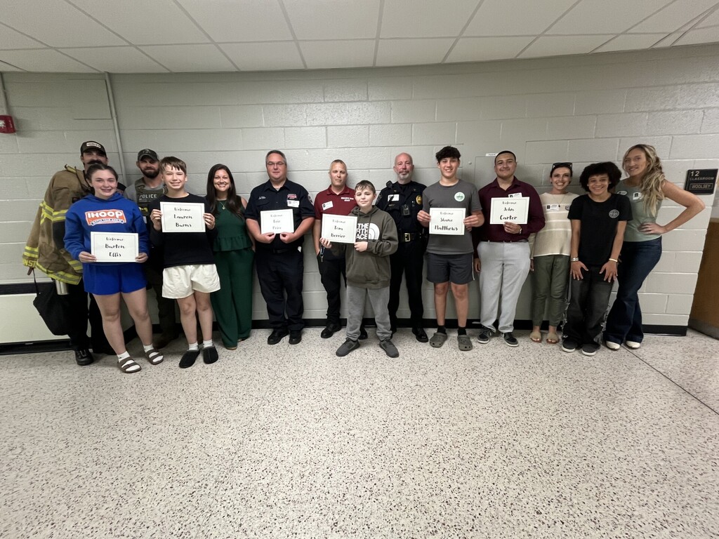 A large group of students and community presenters stand in a school hallway against a light-colored brick wall for a group photo. Several participants in the front row hold printed “Welcome” signs with names on them. The group includes individuals dressed in a variety of professional attire, including uniforms such as firefighting gear and law enforcement clothing, as well as casual and business clothing, representing different careers.