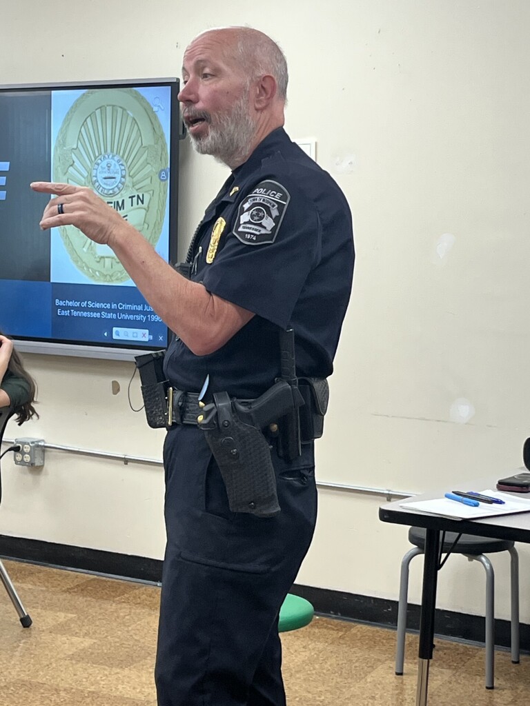 A law enforcement presenter speaks to students in a classroom while standing near a screen displaying a badge graphic and text about education. The presenter wears a uniform with equipment on a duty belt, and a small table with papers and pens sits nearby.