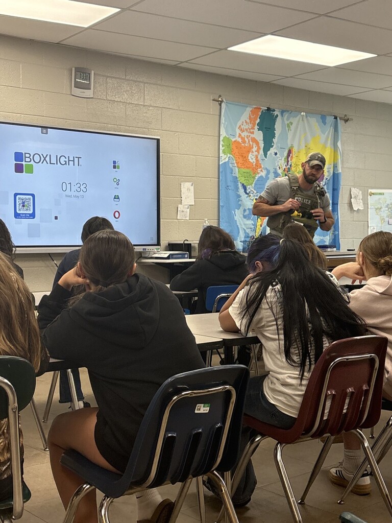 A law enforcement presenter wearing tactical gear demonstrates equipment at the front of a classroom while students sit at desks listening. A digital screen displaying “Boxlight” and a world map poster are visible in the background.