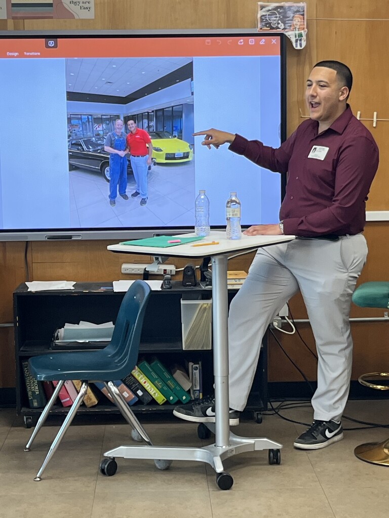 A community presenter stands at a classroom podium gesturing toward a large screen displaying a photo of two people standing next to cars inside a showroom. Papers, water bottles, and classroom materials are visible on the desk in front.