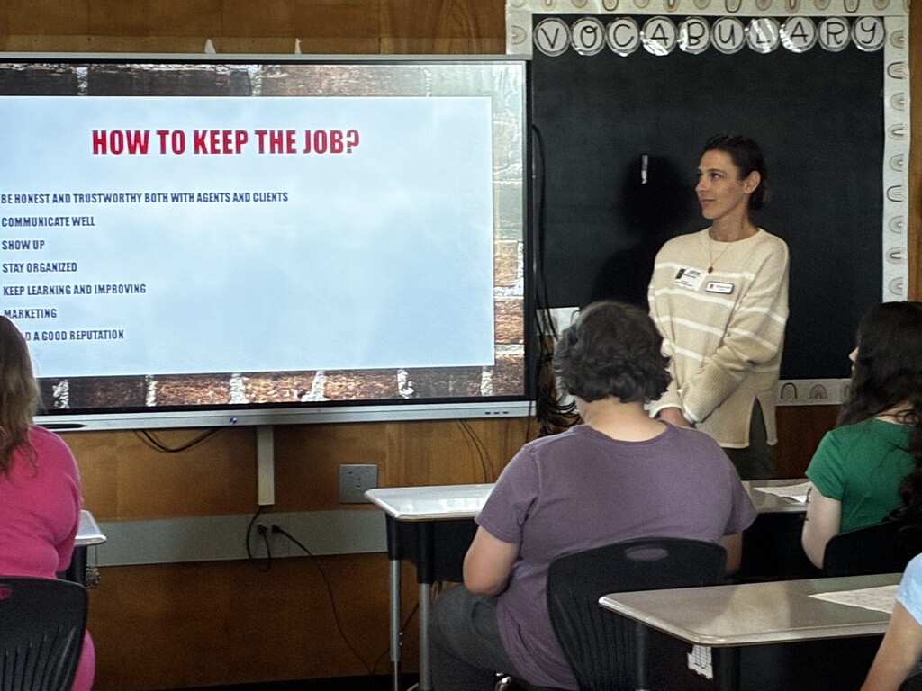 A community presenter stands near a classroom screen displaying a slide titled “How to Keep the Job?” with a list of professional habits. 