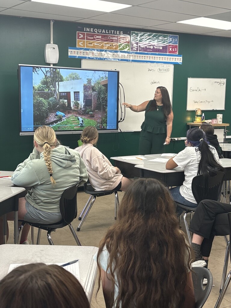 A community presenter stands at the front of a classroom pointing to a large screen displaying a photo of a house and yard, while students sit at desks taking notes. A whiteboard and educational posters are visible on the walls behind.