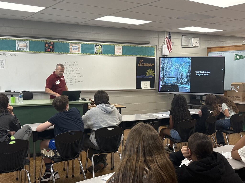 A community presenter stands at the front of a classroom speaking to seated students while using a laptop. A large screen displays a slide reading “Wintertime at Brights Zoo,” and a whiteboard behind shows the daily class schedule.