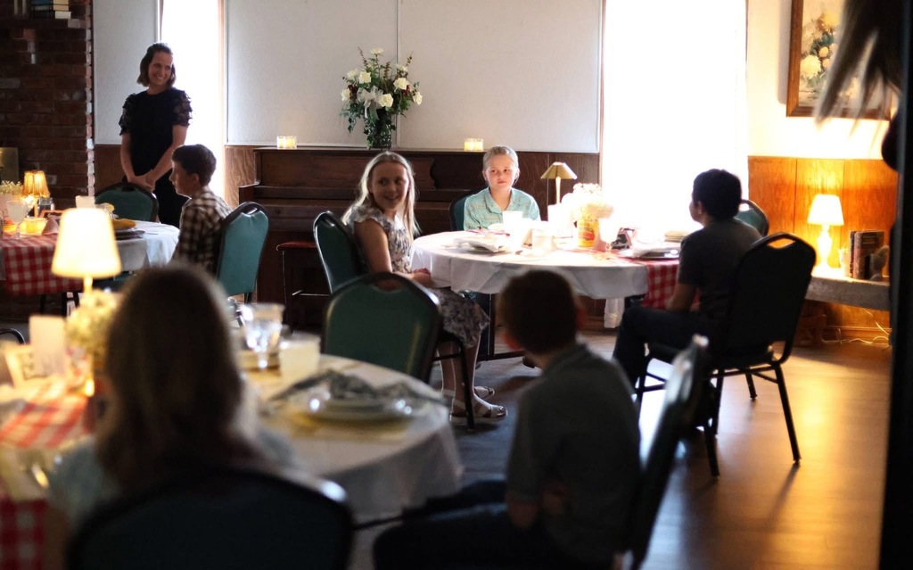 Image of several tables of 5th graders waiting for their meal to be served. A parent volunteer can be seen smiling in the background.