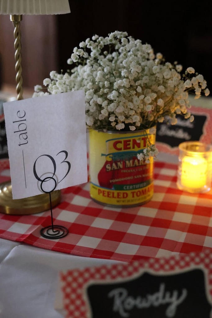 Image of table centerpiece with table number 3 card, tomato can filled with flowers, and a candle on a red checkered table runner