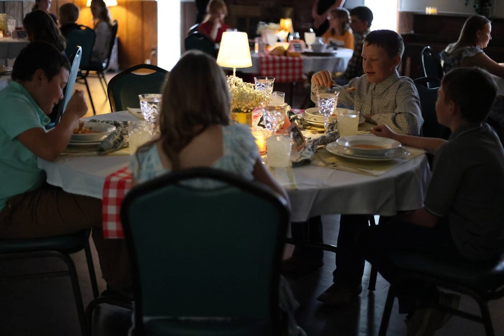 Image of 5th grade students sitting at their table, eating their meal. More tables of students can be seen in the background.