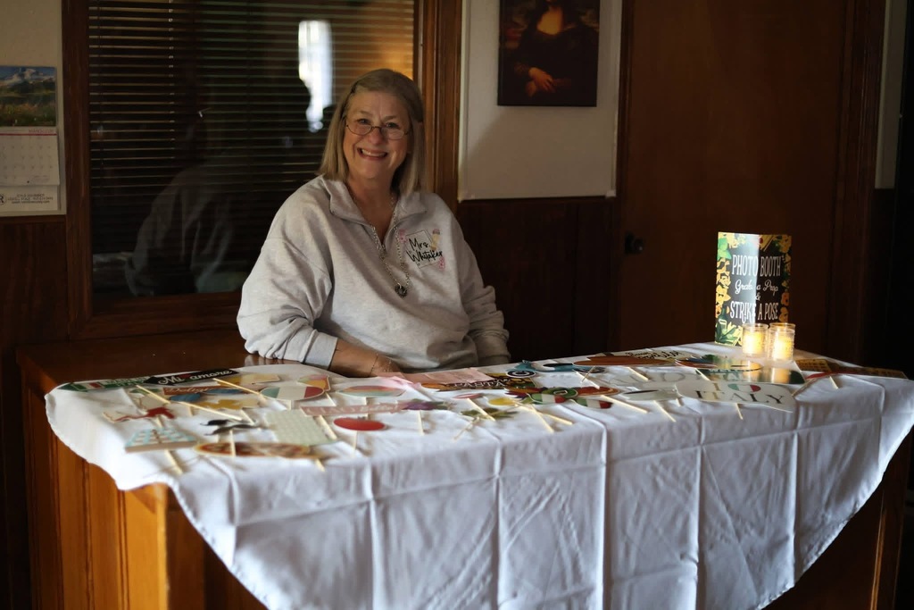 Image of Mrs. Whitaker sitting at a table with a white tablecloth and photo booth props