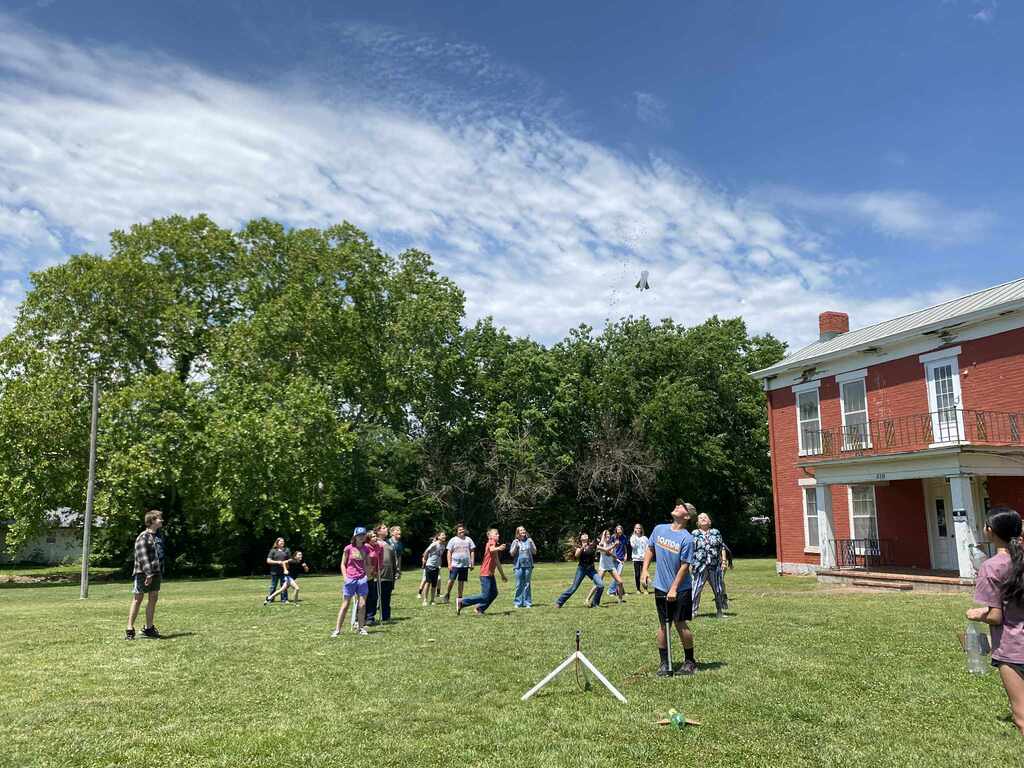 image of students watching a water rocket science demonstration outside with a two-story red house behind them