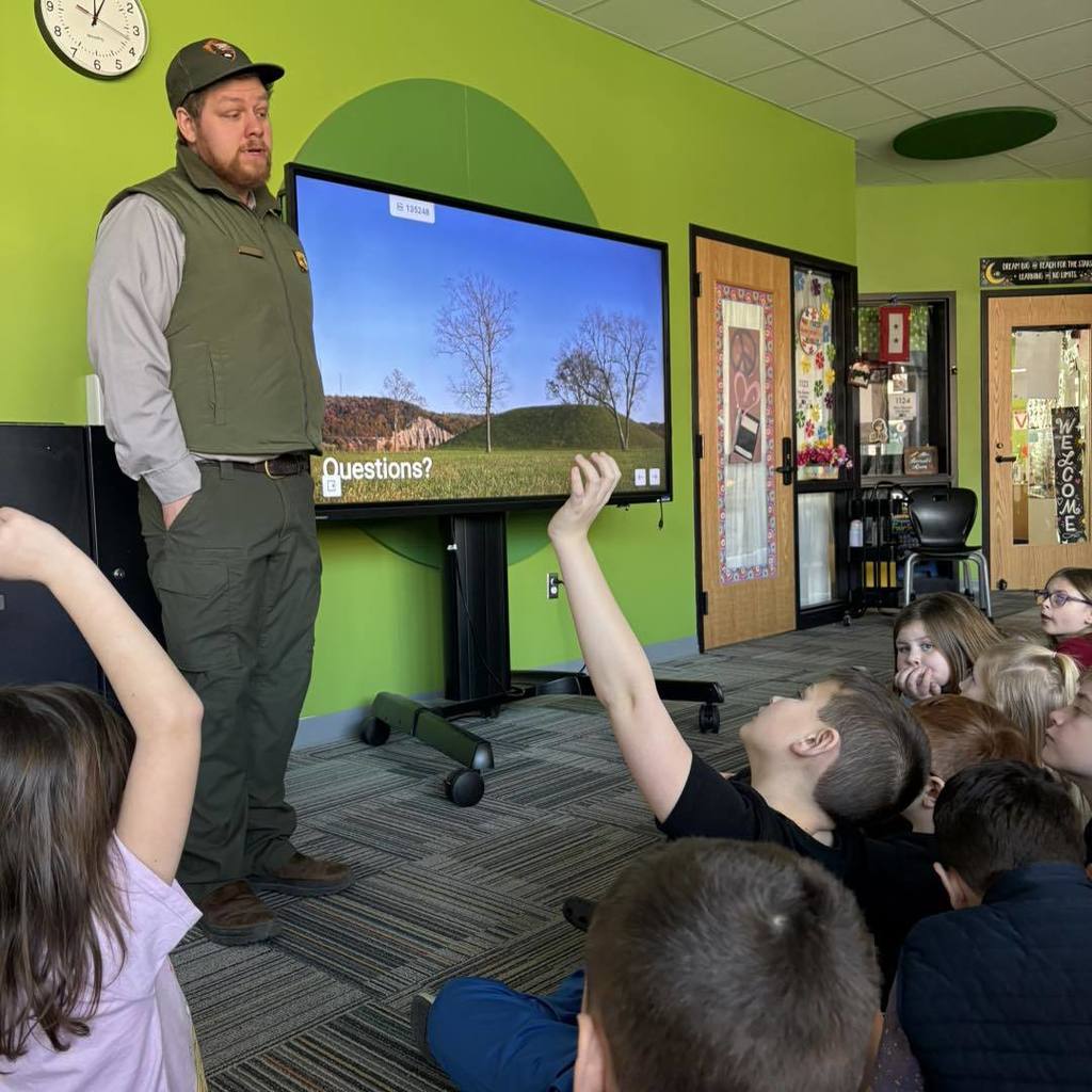 Man stands before class with monitor, showing students an image of a tree on a hill.